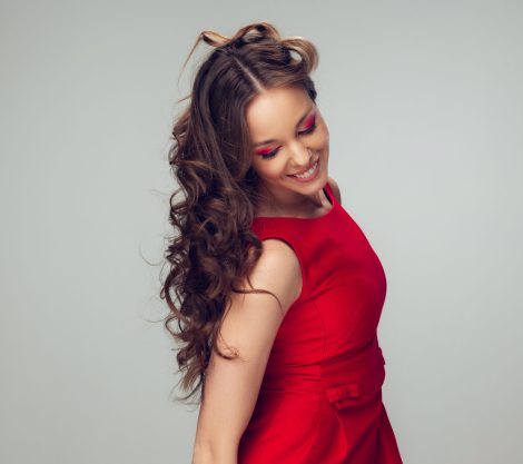 Beautiful young woman with long healthy curly hair and bright make up wearing red dress isolated on grey studio backgroud. Beauty and fashion, hair and skin care, treatment concept. Well-kept girl.