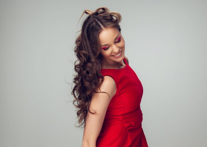 Beautiful young woman with long healthy curly hair and bright make up wearing red dress isolated on grey studio backgroud. Beauty and fashion, hair and skin care, treatment concept. Well-kept girl.
